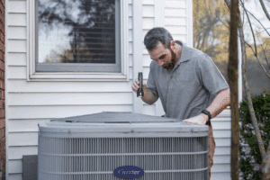 Man inspecting outdoor heat pump unit with handheld flashlight.