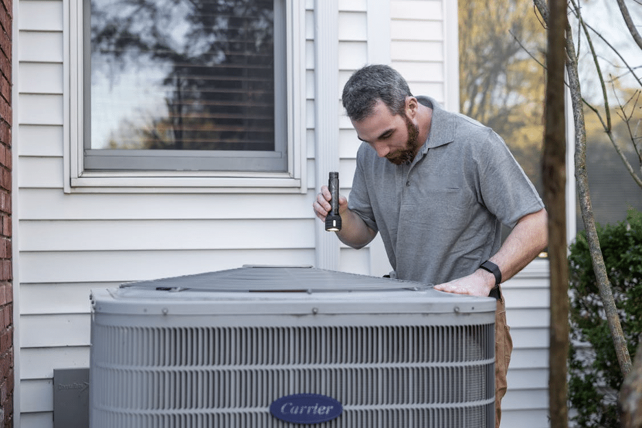 Man inspecting outdoor heat pump unit with handheld flashlight.